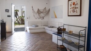 Een moderne woonkamer met een witte bank, een bijzettafeltje en een metalen schapunit in het zwart. Er is een deur die leidt naar een balkon met uitzicht op palmbomen.