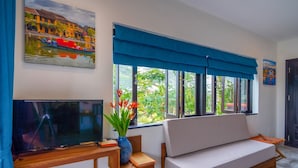 A living room with a TV, a sofa, a window with blue curtains, and a painting on the wall. - Lemongrass Homestay (Hoi An)