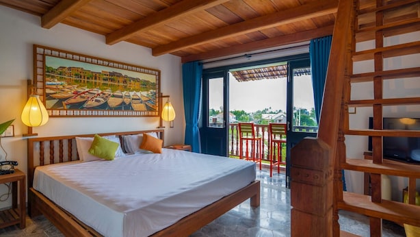 A hotel room with a large bed, wooden ceiling, a painting of boats, a balcony with chairs, and a TV. - Lemongrass Homestay (Hoi An)