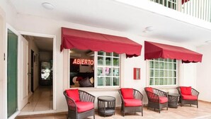 Un espace extérieur aménagé avec des chaises en osier et des coussins rouges, une devanture de magasin avec une enseigne indiquant « OUVERT », et un mur extérieur blanc.
