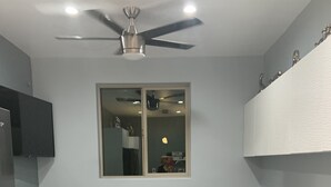 Ein modernes Wohnzimmer mit einer grauen Eckcouch, einem Deckenventilator, einem großen Fenster und einer Küchenzeile mit Fernseher und Schränken.