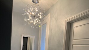 Ein Flur mit einer weißen Tür, einem Spiegel, einem gemusterten Teppich und einer Kandelaberleuchte.