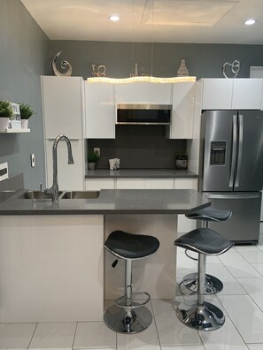 A modern kitchen with a stainless steel refrigerator, a built-in oven, a sink, and two bar stools.