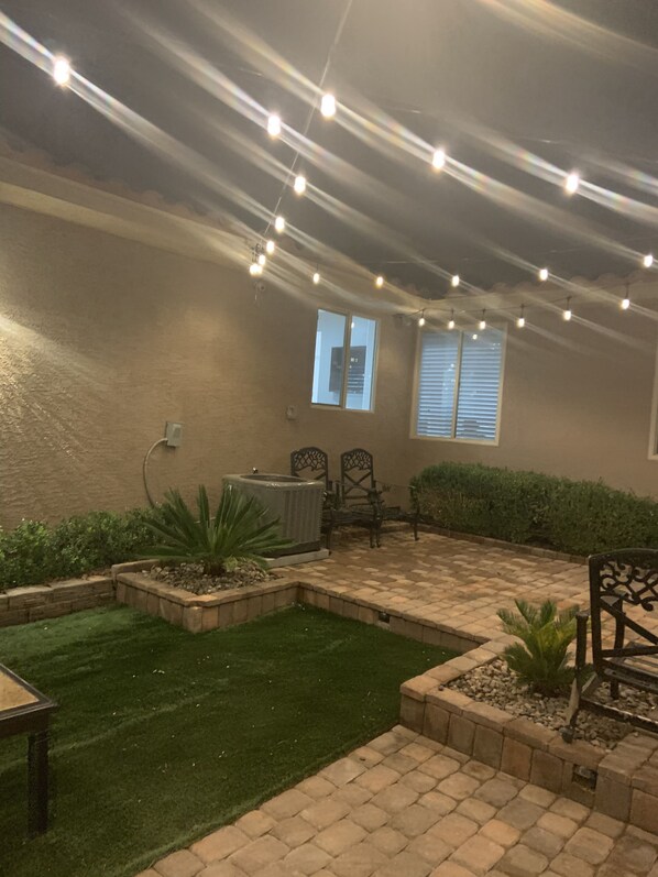 An outdoor patio area with a brick floor, green artificial grass, and a seating arrangement. There are string lights overhead and a small air conditioning unit mounted on the wall.