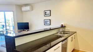 A modern kitchen with a black countertop, white cabinets, a stainless steel sink, and a dishwasher. There is a television on a stand and two framed pictures on the wall. - Coastal Home away from Home (Torquay)