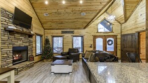 Ein gemütliches Blockhaus-Interieur mit Kamin aus Stein, einem an einer Steinsäule montierten Fernseher, einem Weihnachtsbaum und einem Schild mit der Aufschrift "FARMHOUSE".