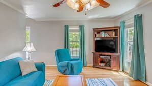 A living room with a teal sofa, a wooden coffee table, a blue armchair, a TV stand with books, and a ceiling fan. - BEACH ISLAND BUM Cozy Cape Code, Pet Friendly Short, Walk to Beach! (Oak Island)