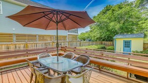 A wooden deck with a glass table and chairs, a brown umbrella, and a small shed in the background. - BEACH ISLAND BUM Cozy Cape Code, Pet Friendly Short, Walk to Beach! (Oak Island)