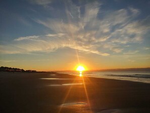 A sunset over a beach with a clear sky and distant buildings. - BEACH ISLAND BUM Cozy Cape Code, Pet Friendly Short, Walk to Beach! (Oak Island)