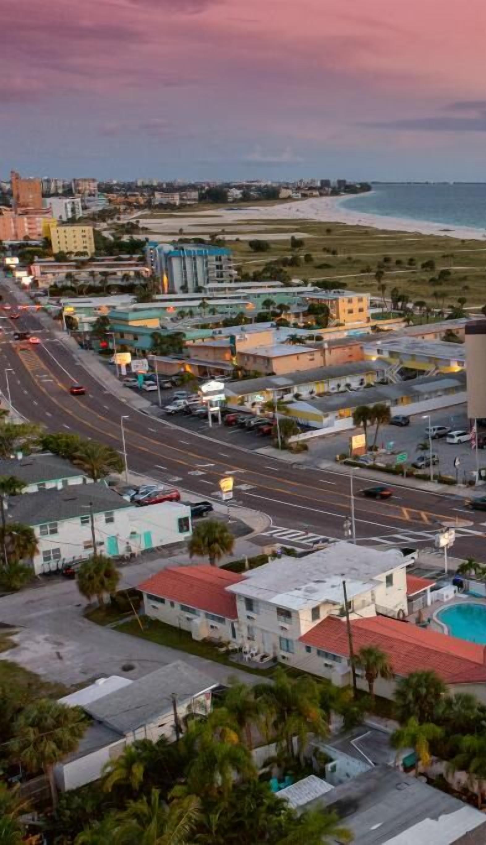 An aerial view of a coastal town with a road leading to a beach, buildings, and a swimming pool.