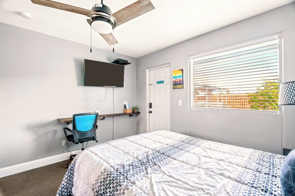 A bedroom with a bed, a desk, a chair, a ceiling fan, a TV, and a window with blinds.