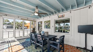 A room with a table, chairs, a ceiling fan, a TV, and a window with a view of palm trees and a building.