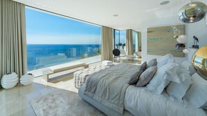 A modern bedroom with a large bed, a view of the ocean, and a balcony with a chair and table.