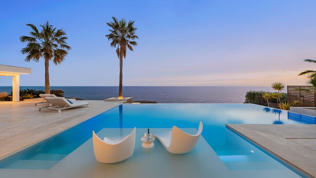 A modern infinity pool with two lounge chairs and a unique seating area, overlooking a beach with palm trees and a clear sky.