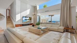 A spacious living room with a large sofa, a coffee table, and a TV mounted on the wall. There is a staircase leading to an upper level, and a view of the ocean through large windows.