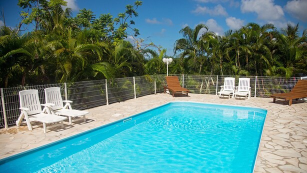 A rectangular swimming pool with clear blue water, surrounded by a white fence. There are white lounge chairs and a wooden bench along the poolside.