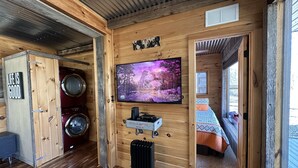 Interior rústico de una cabaña con paredes de madera, un televisor montado, una lavadora y una cama con manta.