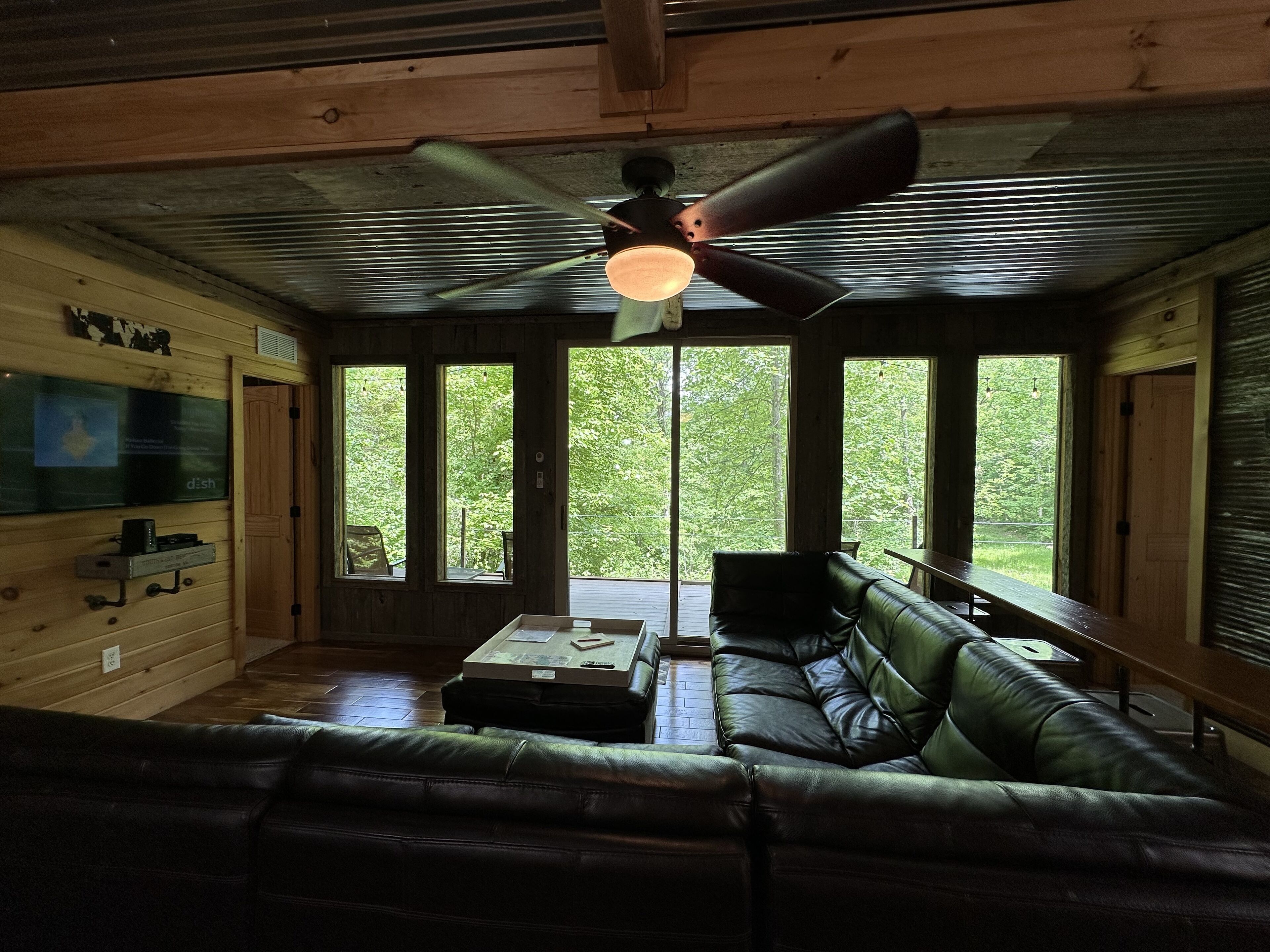 A cozy cabin interior with a leather sofa, a dining table, and a ceiling fan. Large windows offer views of the surrounding forest.