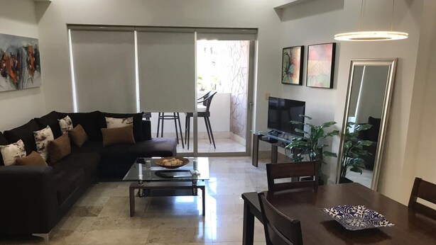Un salon moderne avec un canapé foncé, une table basse en verre, une table à manger en bois avec des chaises, et un téléviseur posé sur un meuble.
