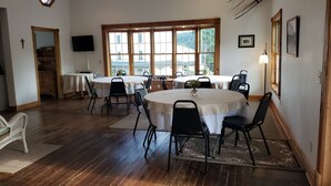 Uma sala de jantar com mesas e cadeiras redondas, janelas amplas, um relógio e uma televisão na parede.