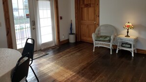 Quarto com piso de madeira, uma cadeira de vime branca, uma mesa e um abajur. Há duas portas, uma com placa de saída e outra com a inscrição 'The Brownwick 102'.