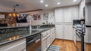 Uma cozinha moderna com bancadas de granito, armários brancos, máquina de lavar louça embutida, fogão e uma grande TV de tela plana fixada na parede.