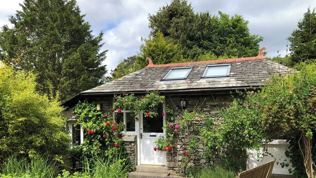 A rustic stone house with a sloped roof, a small porch with a white door, and a garden with various plants and flowers. - A lovely open plan detached cottage in a rural location. (Near Sawrey, near Ambleside)