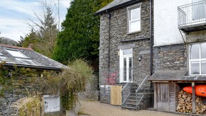 Une maison en pierre avec un escalier en bois menant à une porte, une allée en gravier et une pile de bois de chauffage à l’extérieur.