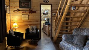 Ein Interieur einer Holzhütte mit Holzbalkendecke, Deckenventilator, Sofa, Sessel, Lampe, Teppich und einer Treppe.