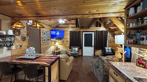 Ein gemütliches Blockhauss-Interieur mit Holzbalkendecke, einem Essbereich mit Tisch und Stühlen, einer Couch und einem an der Wand befestigten Fernseher.