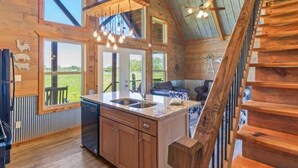 A rustic cabin interior with a kitchen area, wooden staircase, and large windows offering views of the outdoors. - Private Country Getaway (Fitzpatrick)