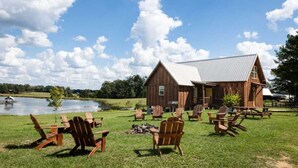 A rustic barn with a metal roof, surrounded by a grassy area with wooden Adirondack chairs and a small fire pit. - Private Country Getaway (Fitzpatrick)