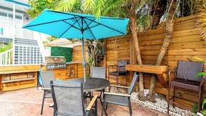 Une terrasse extérieure avec une table ronde, quatre chaises, un parasol bleu, un barbecue et une clôture en bois.