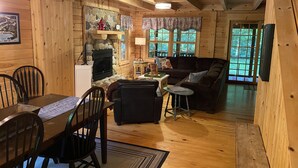 Un interior de cabaña acogedor con comedor, chimenea, sofá y un ventanal grande.