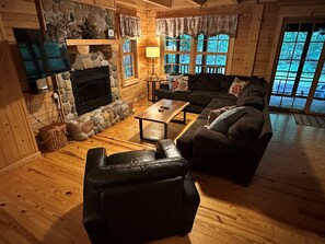 A cozy cabin interior with a stone fireplace, wooden walls, a leather sofa, and a large window offering a view of the outdoors.