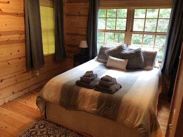 A wooden cabin bedroom with a bed, pillows, a nightstand, and a window with curtains.
