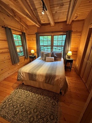 A wooden cabin bedroom with a bed, two bedside lamps, a patterned rug, and a window with curtains.