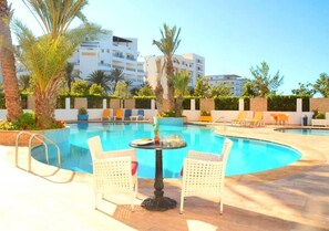 Une piscine avec deux chaises blanches et une table, entourée de palmiers et, au fond, d’un bâtiment blanc.