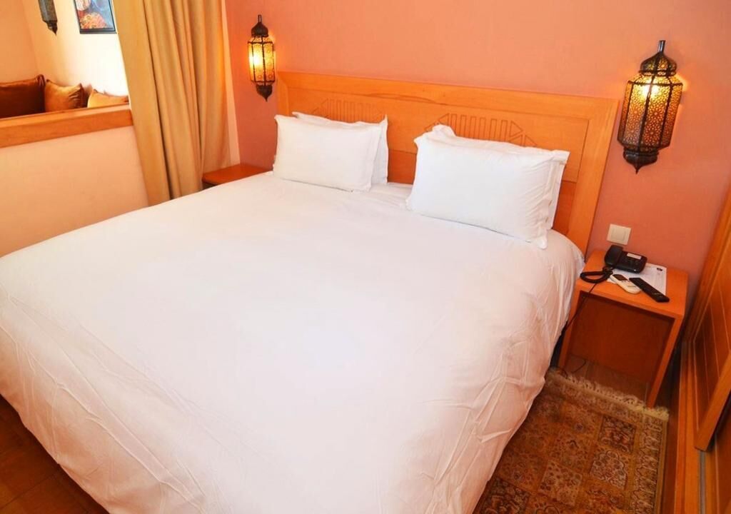 Una cama grande con sábanas blancas, dos lámparas de pared y una mesita de noche con un teléfono y un control remoto.