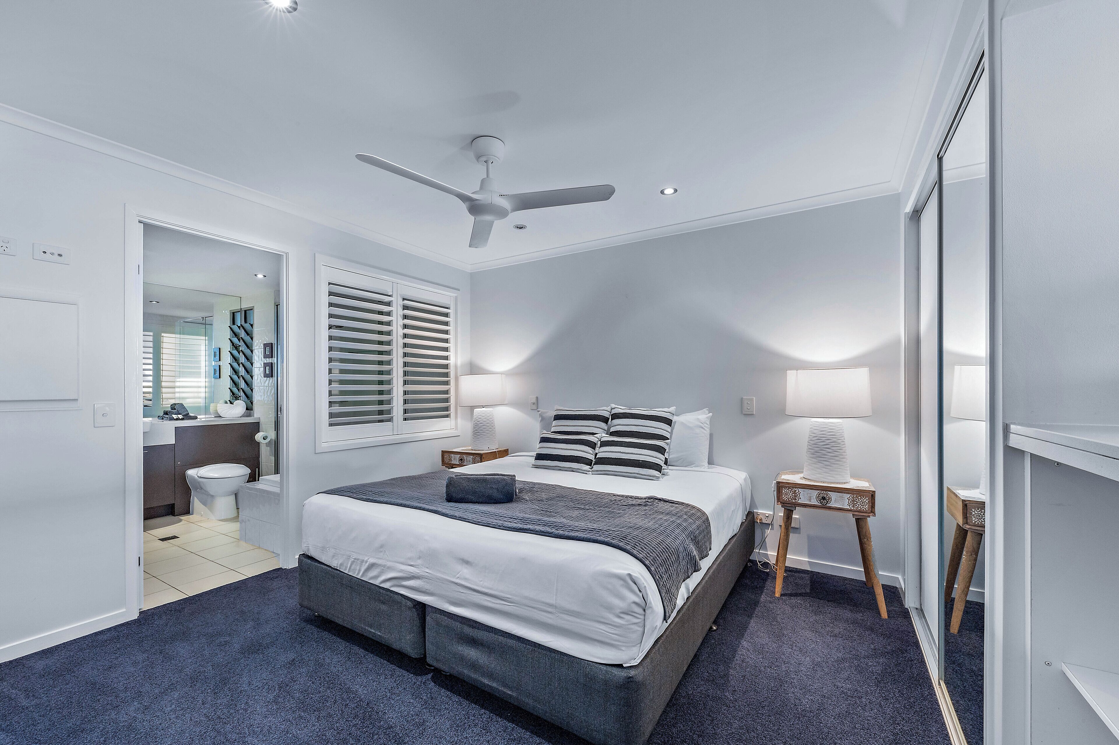 Une chambre à coucher comprenant un lit, des tables de chevet, un ventilateur de plafond et une fenêtre avec des stores.