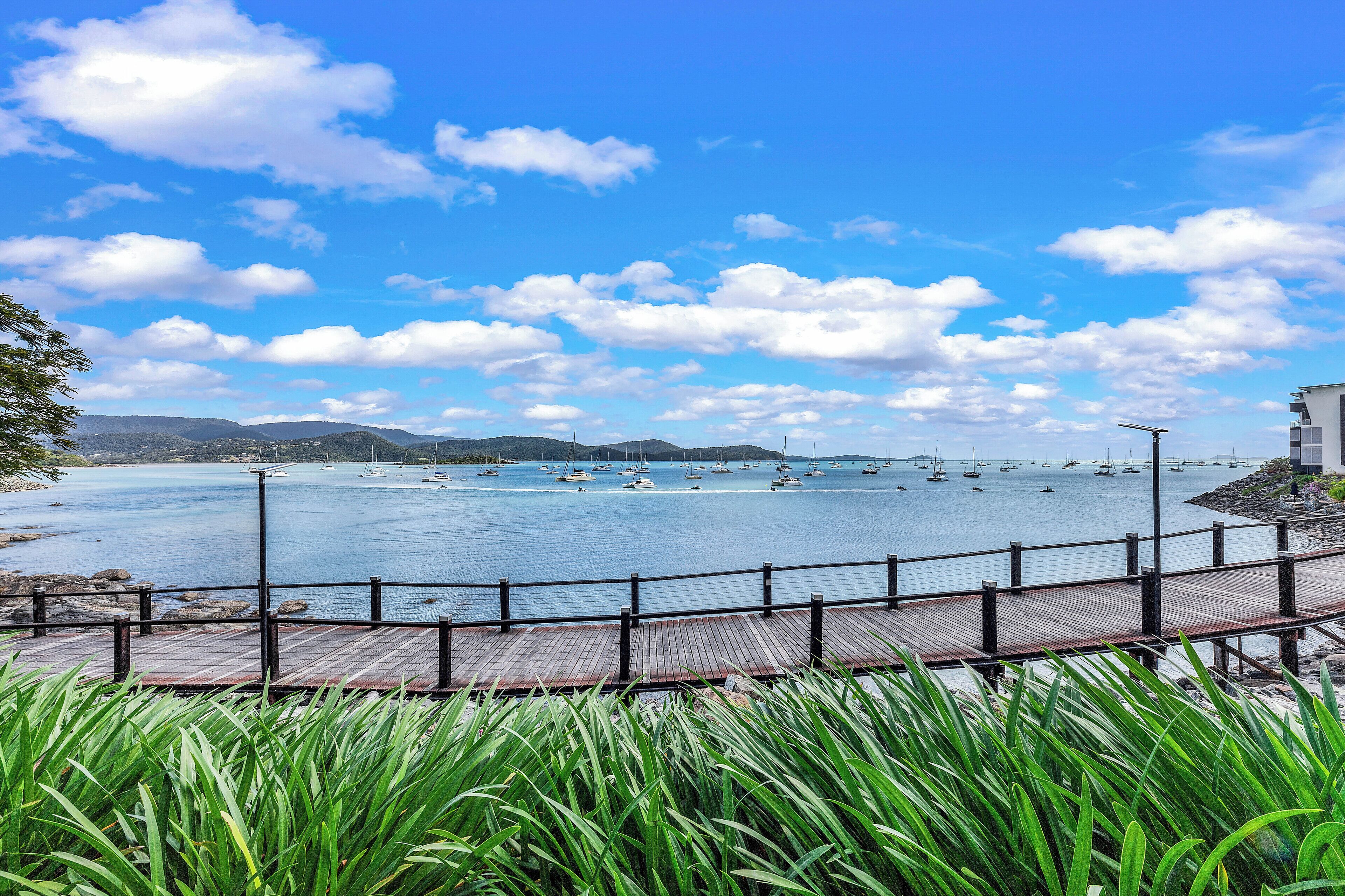 A wooden deck overlooking a calm bay with sailboats, surrounded by greenery and mountains in the distance.