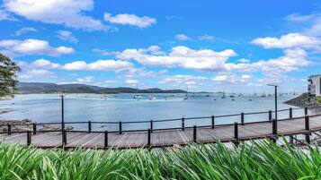 A wooden deck overlooking a calm bay with sailboats, surrounded by greenery and mountains in the distance.