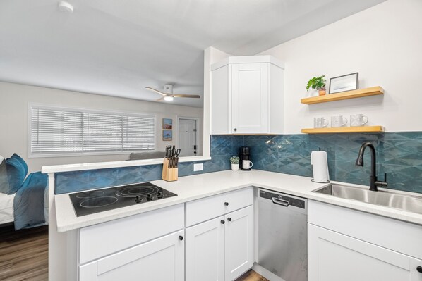 A modern kitchen with white cabinets, a built-in stove, a dishwasher, and a sink. There is a ceiling fan, a window with blinds, and a small shelf with decorative items. - 🌤️ BBR #4, Steps to the Beach, Heated Pool w/Full Kitchen & 2 Beds! (Treasure Island)