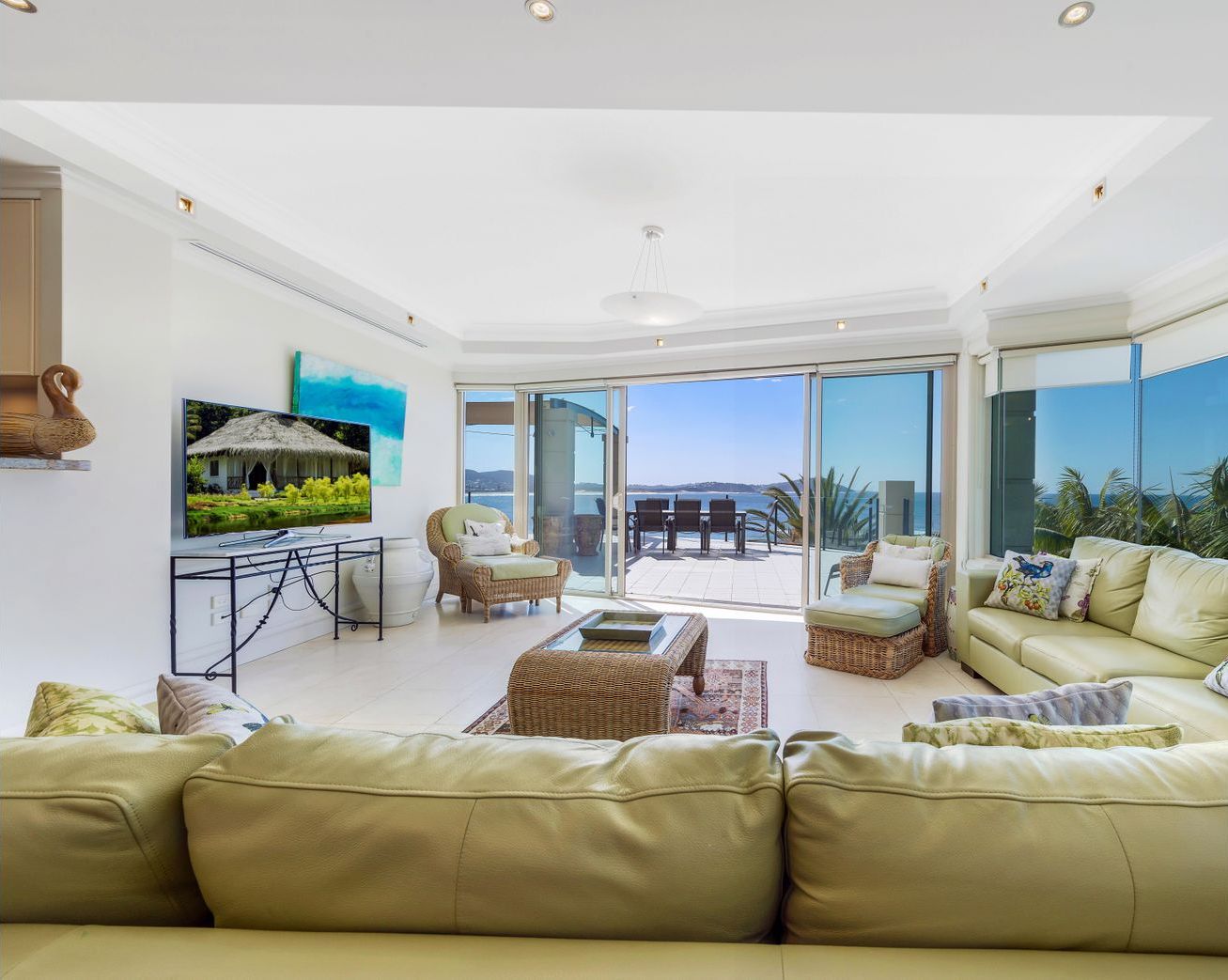 Un salon spacieux avec un grand canapé, une table basse et un téléviseur posé sur un meuble. Il y a un balcon avec vue sur la mer.