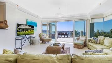A spacious living room with a large sofa, a coffee table, and a TV on a stand. There is a balcony with a view of the sea.