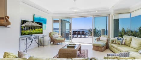 A spacious living room with a large sofa, a coffee table, and a TV on a stand. There is a balcony with a view of the sea.