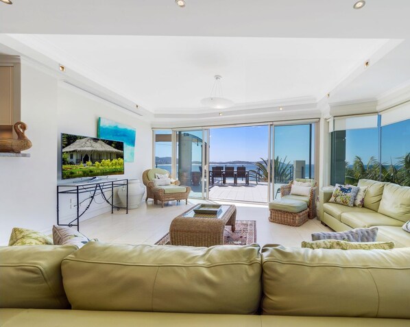 A spacious living room with a large sofa, a coffee table, and a TV on a stand. There is a balcony with a view of the sea.