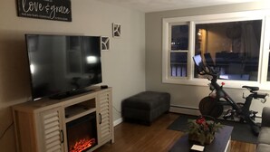 A living room with a flat-screen TV on a wooden cabinet, a fireplace, a couch, a table with a flower vase, and a bicycle leaning against the wall.