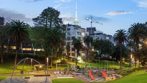 A park with playground equipment, a large tree, and a building with a tower in the background. - *Pool, spa, two balconies, park views!* (Auckland)