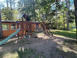 Playhouse with a slide, nestled among trees on a grassy lawn.